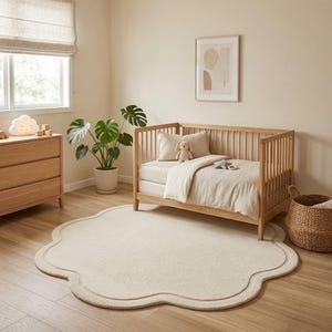May include: A light beige, flower-shaped area rug with a scalloped edge, positioned on a light wood floor in a room featuring a wooden crib with white bedding, a wooden dresser, a potted monstera plant, and a woven basket.