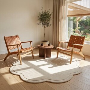 May include: Two light wood armchairs with tan woven leather seats and backrests are positioned on a large, cream-colored, scalloped-edge area rug. Between the chairs sits a small, round, dark brown wooden coffee table with two dark mugs.