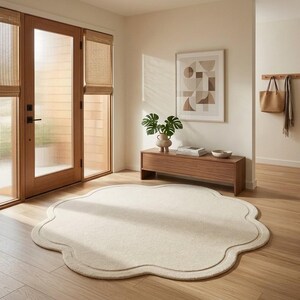 May include: A large, circular, cream-colored area rug with a scalloped, flower-like edge and a subtle embossed border detail. The rug is placed on a light-toned wooden floor in a bright room with a wooden door, a low bench, and wall art.