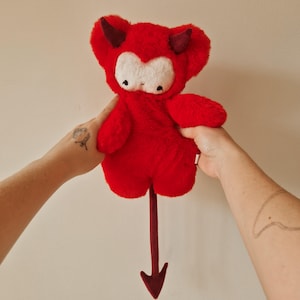 Pode incluir: Um brinquedo de pelúcia em forma de diabo com pelo vermelho brilhante, rosto cor de creme e dois chifres vermelho-escuros. O brinquedo apresenta pequenos olhos pretos, uma boca bordada simples e uma cauda longa e fina vermelho-escura.