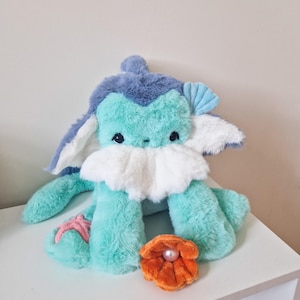 Può includere: Un peluche con morbida pelliccia verde menta, un petto bianco soffice e dettagli blu pervinca. Presenta una piccola stella marina rosa su un arto, un orecchio a forma di conchiglia blu e un accessorio arancione a forma di vongola con una perla.