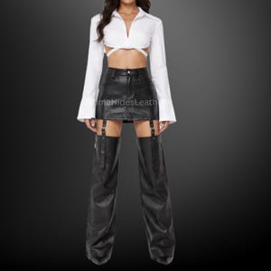May include: A white long-sleeved cropped shirt with a collar and wrap-around waist ties, paired with a black leather mini skirt and matching black leather leg warmers that attach to the skirt with vertical buckled straps and metal hardware.