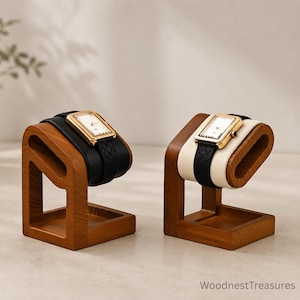 May include: Two wooden watch stands with rectangular bases and angled supports. One stand holds a gold-tone rectangular watch with a black textured strap on a black cushion. The other holds a gold-tone watch on a cream cushion. Text reads WoodnestTreasures.
