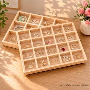 May include: Two light-colored wooden jewelry organizer trays with multiple square compartments. The trays contain various gold-toned rings, earrings, and hair accessories. The text WoodnestTreasures is printed in the bottom right corner.