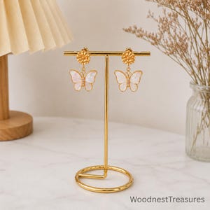 May include: A pair of gold-toned earrings hanging on a T-shaped jewelry stand. Each earring features a textured circular stud connected to a white, iridescent butterfly-shaped charm with a central vertical row of small, clear, sparkling stones.