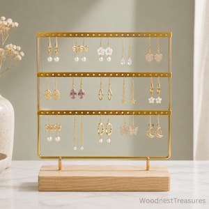 May include: A gold metal earring display stand with three horizontal bars featuring evenly spaced holes for hanging jewelry. The stand is mounted on a rectangular light-colored wooden base. The text WoodnestTreasures is visible in the bottom right.
