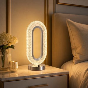 May include: A modern oval-shaped table lamp with a textured, crystalline outer frame that emits a warm, soft glow. The lamp features a silver-toned circular base and a hollow centre, providing a sleek, illuminated accent for a bedside table.