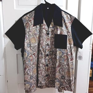May include: A short-sleeved button-down shirt with a black collar, sleeves, and pocket. The main body of the shirt features a patterned design with various illustrations in shades of beige, blue, and red. The shirt is hanging on a black hanger.