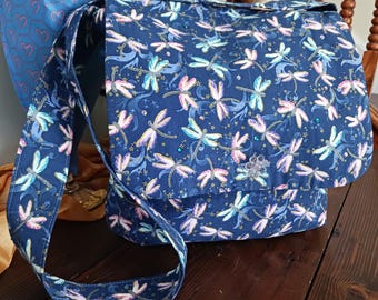 May include: A navy blue messenger bag with a dragonfly pattern in shades of pink, blue, and white. The bag has a flap closure and a long shoulder strap. A decorative embellishment is on the flap.