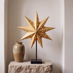 May include: A nine-pointed star-shaped lamp shade in a light tan color with cut-out star patterns. The lamp shade is mounted on a thin black metal rod with a square black base, positioned next to a smooth, light brown ceramic vase.