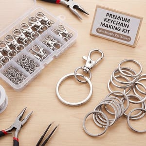 May include: A silver keychain making kit featuring a clear plastic organizer box filled with metal lobster clasps and jump rings, a set of pliers, tweezers, and a sign labeled PREMIUM KEYCHAIN MAKING KIT Sam Supply Co (Silver) with loose rings.