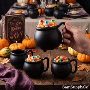 May include: A matte black ceramic cauldron mug with a rounded body and a single handle, filled with colorful gummy candies shaped like eyeballs and assorted sweets. The mug features a wide rim and small feet at the base, designed for holding beverages.