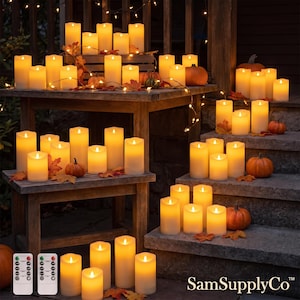 May include: A collection of cylindrical LED pillar candles with a warm yellow glow, arranged on wooden steps and a table alongside small orange pumpkins and autumn leaves. Two white remote controls with multiple buttons are visible in the foreground.