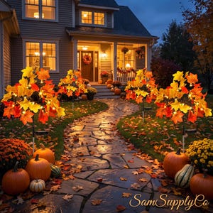 May include: Four illuminated artificial maple trees with vibrant orange, yellow, and red leaves lining a stone walkway. Each tree features small warm white lights nestled among the foliage, accompanied by orange pumpkins and yellow mums.
