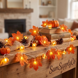 May include: A string of LED lights featuring orange maple leaves and small pumpkins. The lights are draped over two stacked books, pinecones, and a burlap ribbon. The text SamSupplyCo is visible on the wooden surface in the foreground.