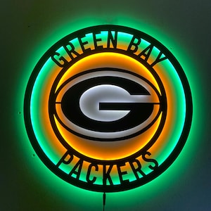 May include: A circular Green Bay Packers wall sign featuring a black frame with the text GREEN BAY PACKERS and a central G logo. The sign is illuminated with a bright green outer ring and a warm yellow inner glow, creating a high-contrast display.