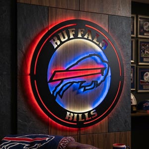 Puede incluir: Un letrero circular de pared con el logotipo de los Buffalo Bills en metal negro con retroiluminación LED roja y azul. La palabra BUFFALO está arqueada en la parte superior y BILLS en la inferior en letras metálicas sobre un fondo de madera.