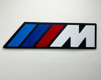 May include: A rectangular wall sign featuring the letter M in white next to three diagonal stripes in red, dark blue, and light blue. The design is set against a black frame with visible mounting screws at the corners, creating a bold graphic display.