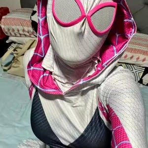 May include: A Spider-Gwen cosplay costume featuring a white and black bodysuit with pink accents. The hood and mask have pink trim and large, white-mesh eye coverings. The costume has a spiderweb design.