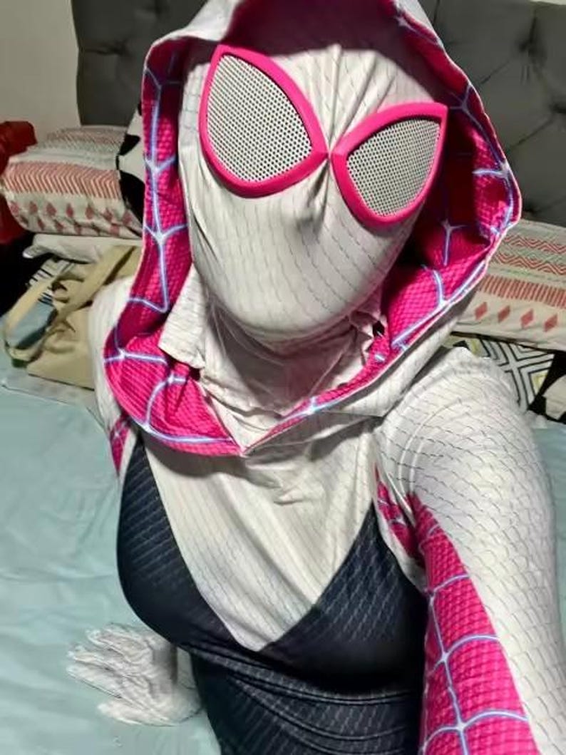 May include: A Spider-Gwen cosplay costume featuring a white and black bodysuit with pink accents. The hood and mask have pink trim and large, white-mesh eye coverings. The costume has a spiderweb design.