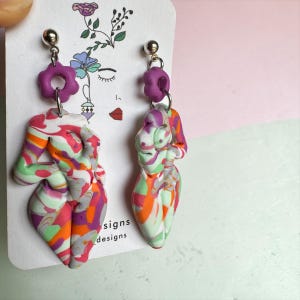 Puede incluir: Un par de pendientes hechos a mano con colgantes abstractos de forma femenina. Los colgantes son multicolores con remolinos de naranja, rosa, verde y blanco. Cada pendiente tiene una flor morada y un perno plateado.