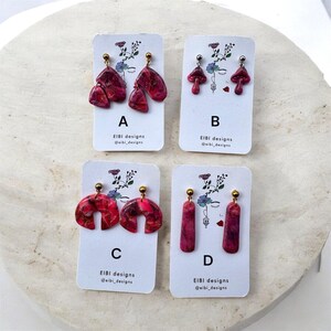 Puede incluir: Se muestran cuatro pares de pendientes hechos a mano. Los estilos incluyen formas abstractas, diseños de setas y barras rectangulares. Cada par es una vibrante mezcla de tonos rojos y rosas, con postes dorados. Las tarjetas muestran el texto "EIBI designs @eibi_designs".