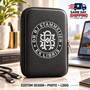 May include: A black rectangular zippered case featuring a white circular emblem on the front. The emblem contains the text DR RJ STAMMEIJER and EX LIBRIS surrounding an intricate monogram of the letters R, J, S, and B in a stylized font.