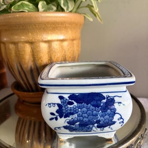 May include: A square ceramic planter with a white base and blue hand-painted grape vine motifs. The planter features a flared rim with a thin blue border and rests on four small feet. The design includes clusters of grapes and leaves.