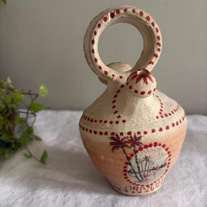 May include: A beige terracotta vessel with a circular handle and a small spout. The surface features hand-painted red dots and a circular illustration of palm trees. The word ORAN is painted in red near the base of the rounded ceramic body.