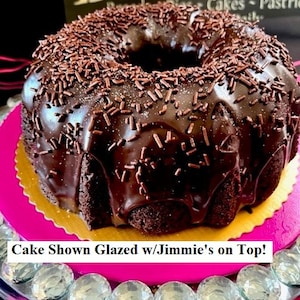 May include: A chocolate cake with a chocolate glaze and sprinkles on top. The cake is on a pink plate with a gold rim. The cake is in front of a sign that says "Baker".
