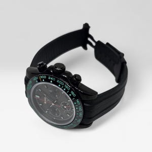 May include: A black chronograph wristwatch featuring a dark dial with three sub-dials and a tachymeter bezel with green numerals. The watch has a black case and a textured black rubber strap with a deployment clasp, showing a clear side profile.