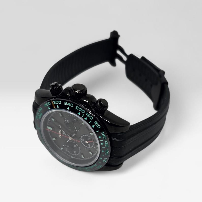 May include: A black chronograph wristwatch featuring a dark dial with three sub-dials and a tachymeter bezel with green numerals. The watch has a black case and a textured black rubber strap with a deployment clasp, showing a clear side profile.