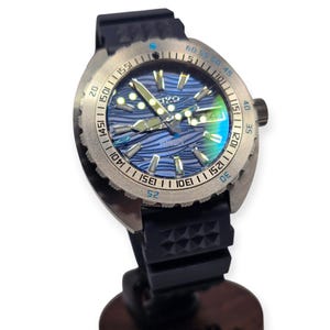 May include: A stainless steel wristwatch with a blue wave-patterned dial. The watch has a black rubber strap and a rotating bezel with minute markers. The watch face displays the brand name and the date.