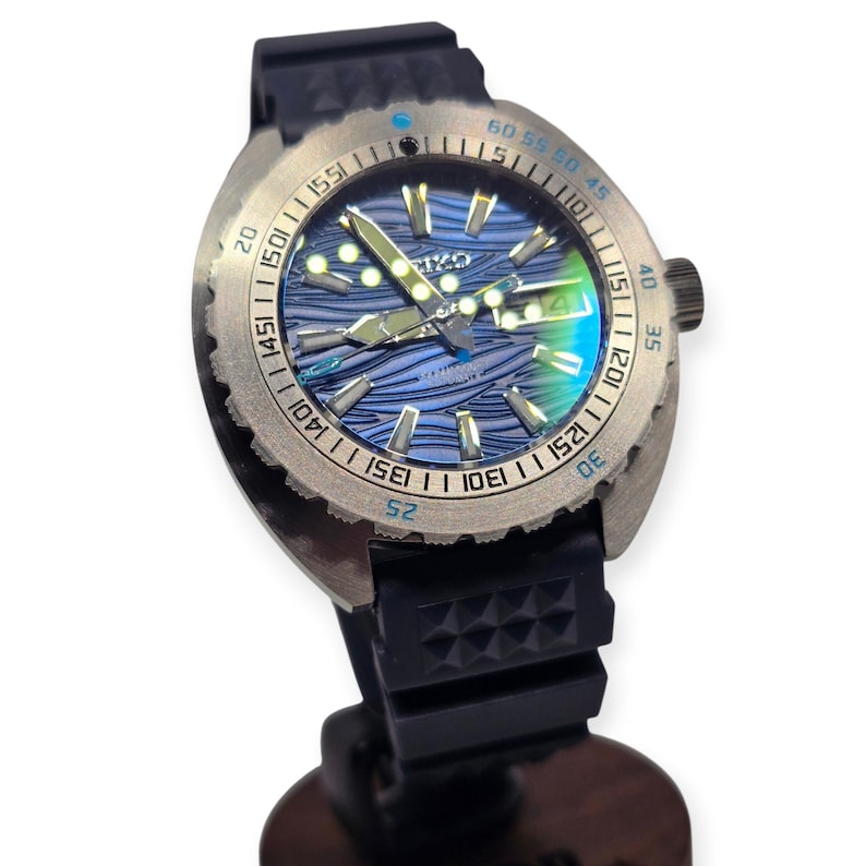May include: A stainless steel wristwatch with a blue wave-patterned dial. The watch has a black rubber strap and a rotating bezel with minute markers. The watch face displays the brand name and the date.