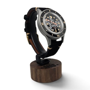 May include: A Seiko automatic wristwatch with a black dial featuring a grid pattern, gold-tone indices, and a black rotating bezel with gold numerals. The watch has a black leather strap with tan stitching and rests on a cylindrical wooden stand.