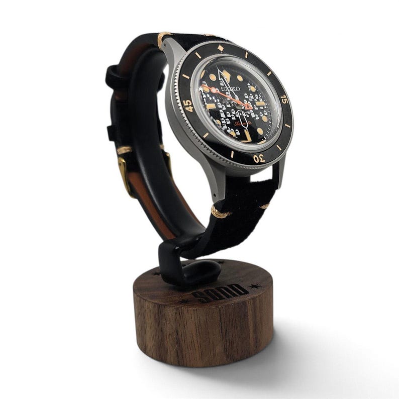 May include: A Seiko automatic wristwatch with a black dial featuring a grid pattern, gold-tone indices, and a black rotating bezel with gold numerals. The watch has a black leather strap with tan stitching and rests on a cylindrical wooden stand.