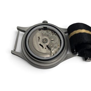 May include: A stainless steel watch case with the back removed to reveal the internal mechanical movement. The watch features a matte grey finish and is attached to a black and tan striped nylon strap with a metal buckle and spring bars.