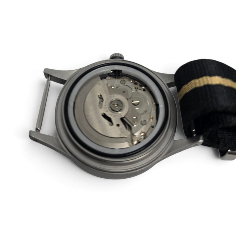 May include: A stainless steel watch case with the back removed to reveal the internal mechanical movement. The watch features a matte grey finish and is attached to a black and tan striped nylon strap with a metal buckle and spring bars.
