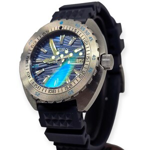 May include: A stainless steel wristwatch with a blue and silver face and a black rubber band. The watch face has a wave-like pattern and luminous hands. The bezel has minute markers. The date is displayed at the 3 o'clock position.