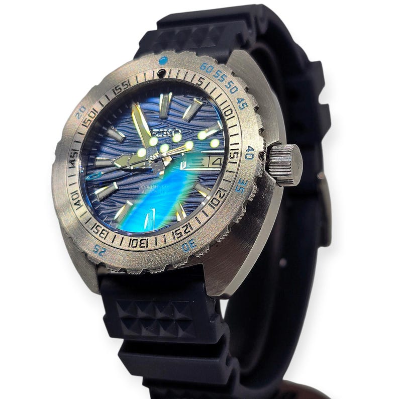 May include: A stainless steel wristwatch with a blue and silver face and a black rubber band. The watch face has a wave-like pattern and luminous hands. The bezel has minute markers. The date is displayed at the 3 o'clock position.