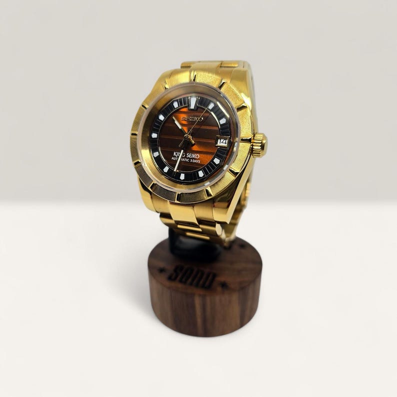 Op de afbeelding: Een goudkleurig Seiko King Seiko automatisch polshorloge met een bruine en zwarte wijzerplaat. Het horloge heeft een gouden armband en wordt getoond op een houten standaard. De wijzerplaat heeft de tekst "King Seiko Automatic 1504S".