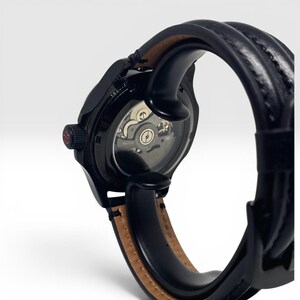 May include: A black wristwatch featuring an exhibition case back that reveals the internal mechanical movement. The watch has a black textured strap with a tan leather lining and a black crown with a small red accent on the side.