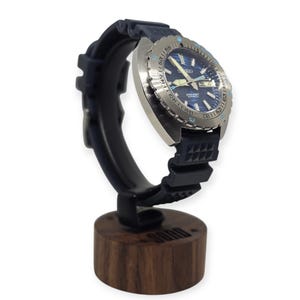 May include: A stainless steel Seiko automatic wristwatch with a blue dial and a black rubber strap is displayed on a black watch stand with a wooden base. The watch has a rotating bezel and is designed for diving.
