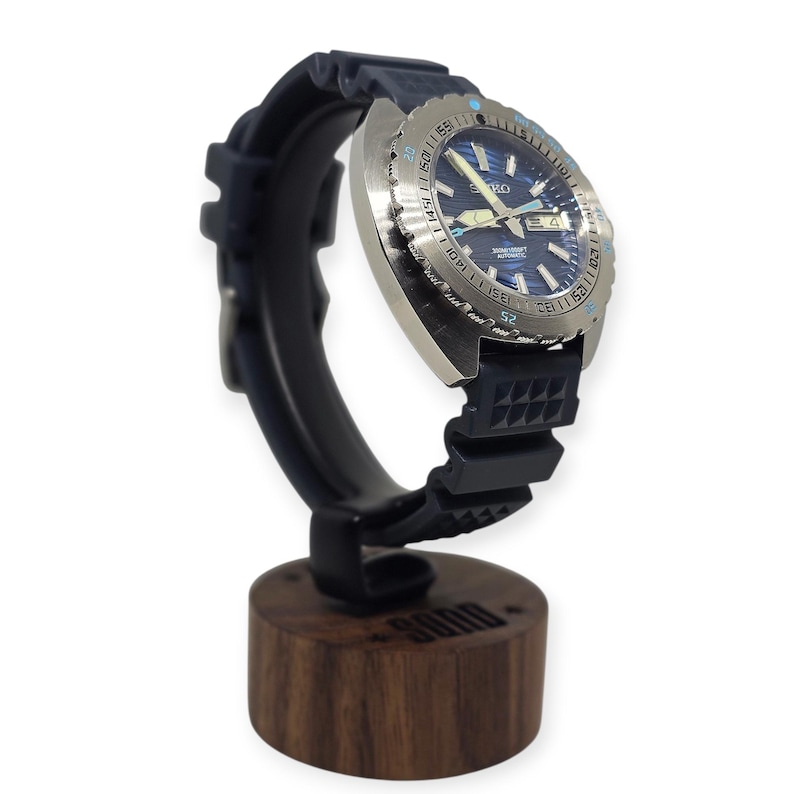May include: A stainless steel Seiko automatic wristwatch with a blue dial and a black rubber strap is displayed on a black watch stand with a wooden base. The watch has a rotating bezel and is designed for diving.