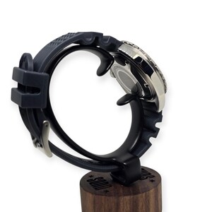 May include: A stainless steel wristwatch with a dark blue rubber strap is displayed on a black stand. The watch has a silver bezel and a dark face. The stand is mounted on a wooden base with text.