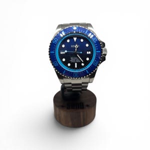 May include: A stainless steel wristwatch with a blue and black rotating bezel, a black dial featuring luminous hour markers, and the text Sea-Dweller 12800ft=3900m Superlative Chronometer Officially Certified. The watch has a metal link bracelet.