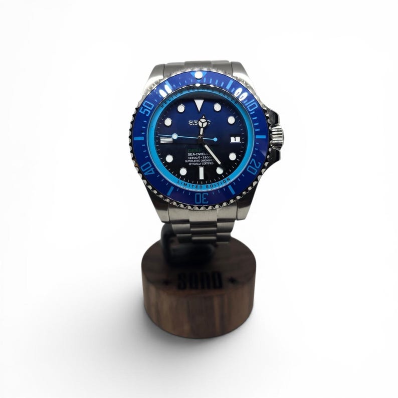 May include: A stainless steel wristwatch with a blue and black rotating bezel, a black dial featuring luminous hour markers, and the text Sea-Dweller 12800ft=3900m Superlative Chronometer Officially Certified. The watch has a metal link bracelet.