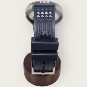 May include: A close-up of a wristwatch with a dark blue band and a silver-colored watch face. The band features a geometric pattern of triangles. The watch is displayed on a wooden stand.