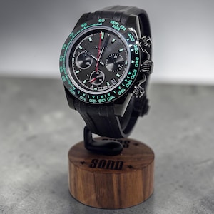 May include: A black chronograph wristwatch with a dark grey dial, three sub-dials, and a tachymeter bezel featuring green luminous markings. The watch has a black silicone strap and is mounted on a cylindrical wooden display stand with the text SQND.