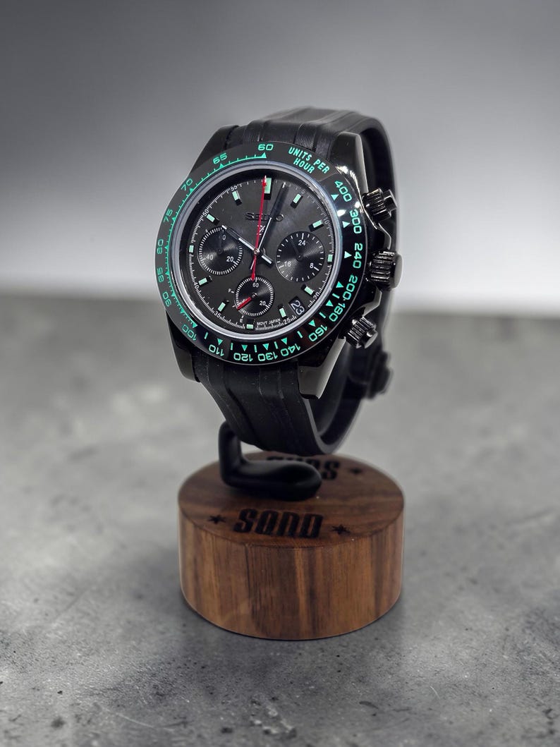 May include: A black chronograph wristwatch with a dark grey dial, three sub-dials, and a tachymeter bezel featuring green luminous markings. The watch has a black silicone strap and is mounted on a cylindrical wooden display stand with the text SQND.